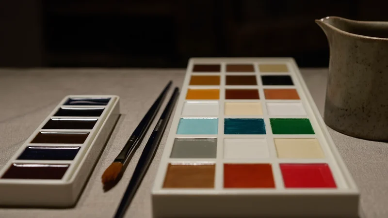 Aquarellfarben in Näpfchen, Haarpinsel und Keramik-Wasserglas auf hellem Leinentuch